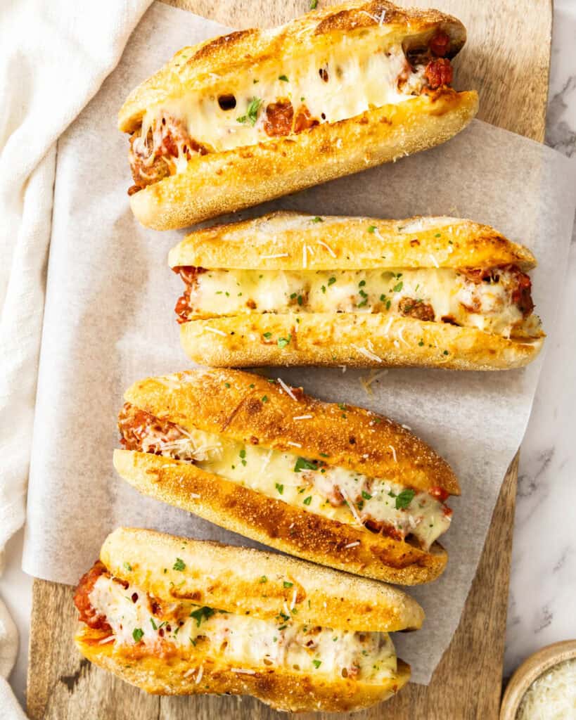 An overhead view of four toasted meatball subs on parchment paper over a cutting bvoard.