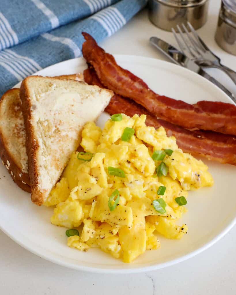 A close up view of microwave scrambled eggs garnished with thin sliced green onions on a plate with bacon and toast. 