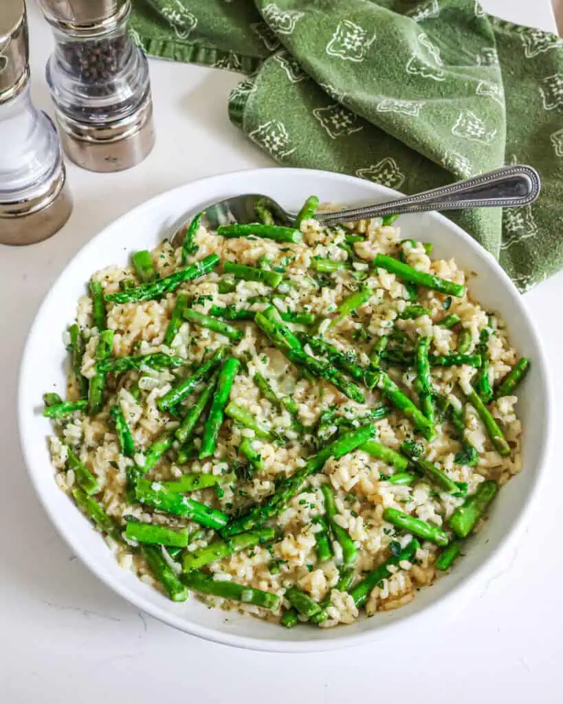 Asparagus risotto in a serving bowl with a metal spoon. There is a salt shaker, pepper shaker, and green kitchen towel in the background. 