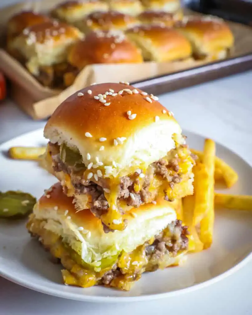 Two stacked cheeseburger sliders on a single serving plate with fries and pickles. 