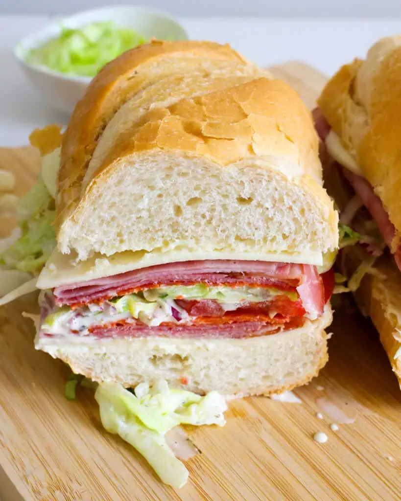 Some of the steps to making Italian grinder sandwiches. A close up view of a grinder sandwich with provolone cheese, smoked ham, pepperoni, Genoa salami, tomatoes, and a tangy mayonnaise iceberg salad all on soft Italian bread.