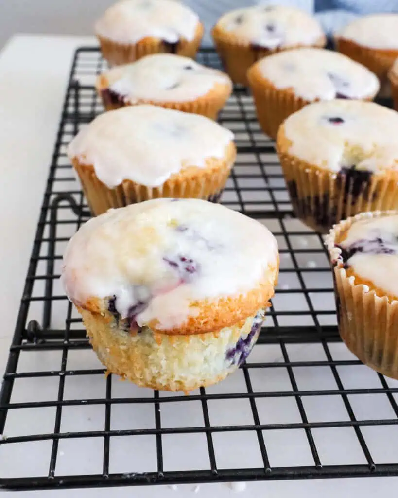 A lemon blueberry muffin with the paper liner rmoved on a wire cooking rack with other lemon blueberry muffins. 