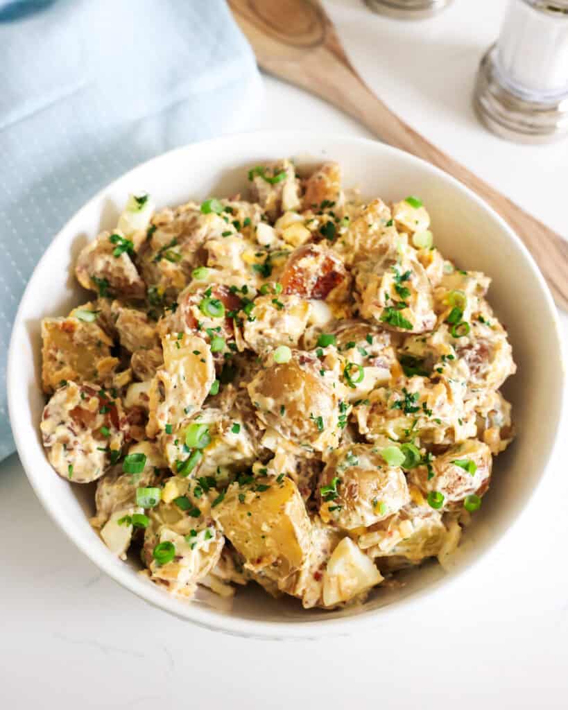 An overhead view of a large bowl of loaded baked potato salad with a wooden spoon and salt and pepper shaker in the background. 