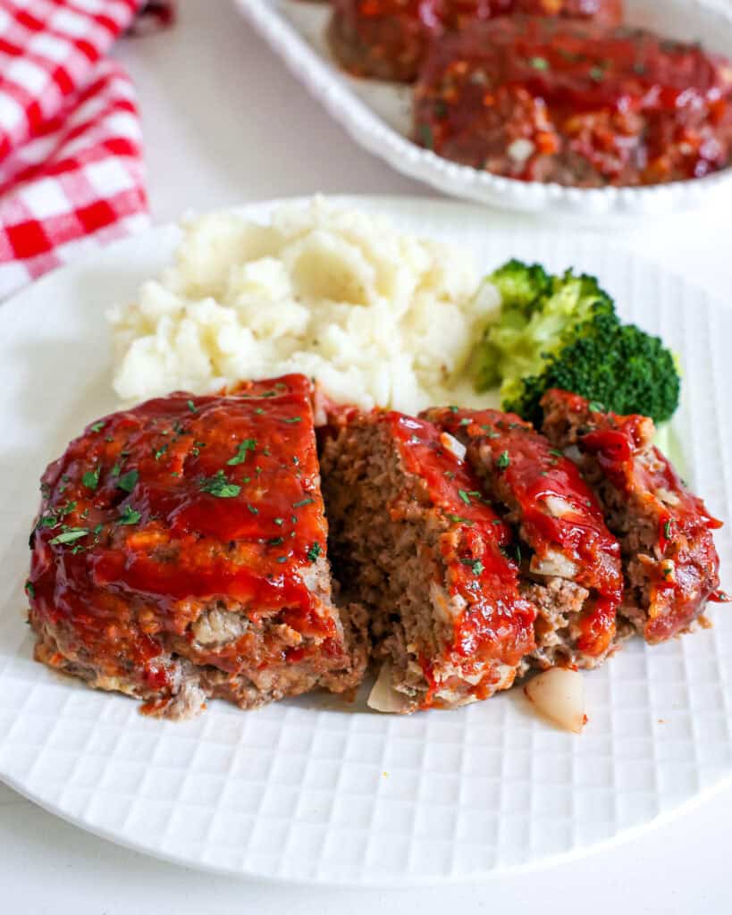 A white serving plate with a mini meatloaf, mashed potatoes, and broccoli with a platter full of mini meatloaves in the background. 
