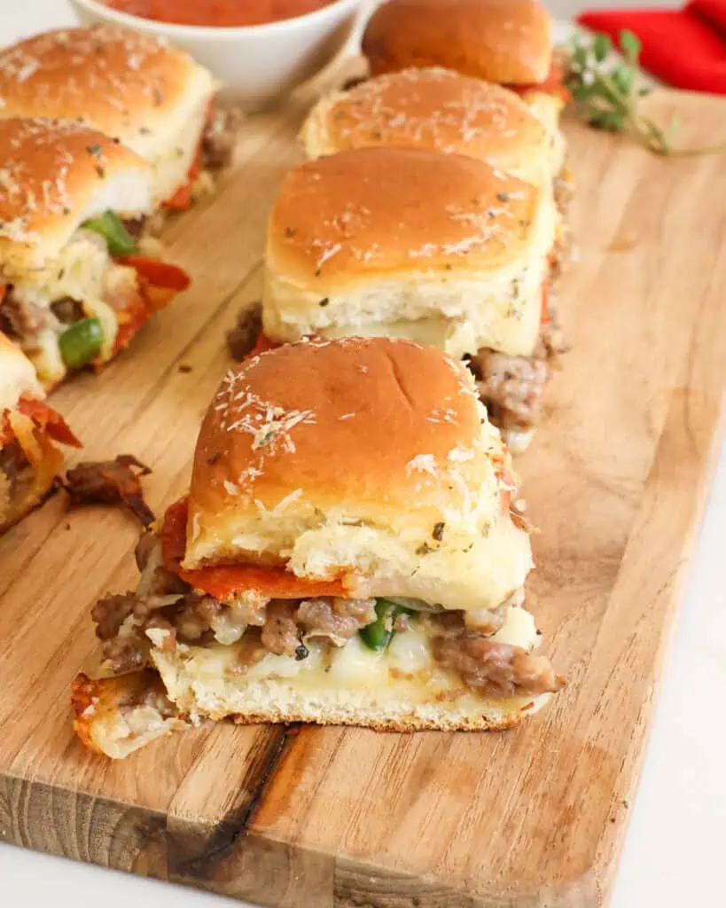Separated pizza sliders on a wooden cutting board with a bowl of pizza sauce in the background. 