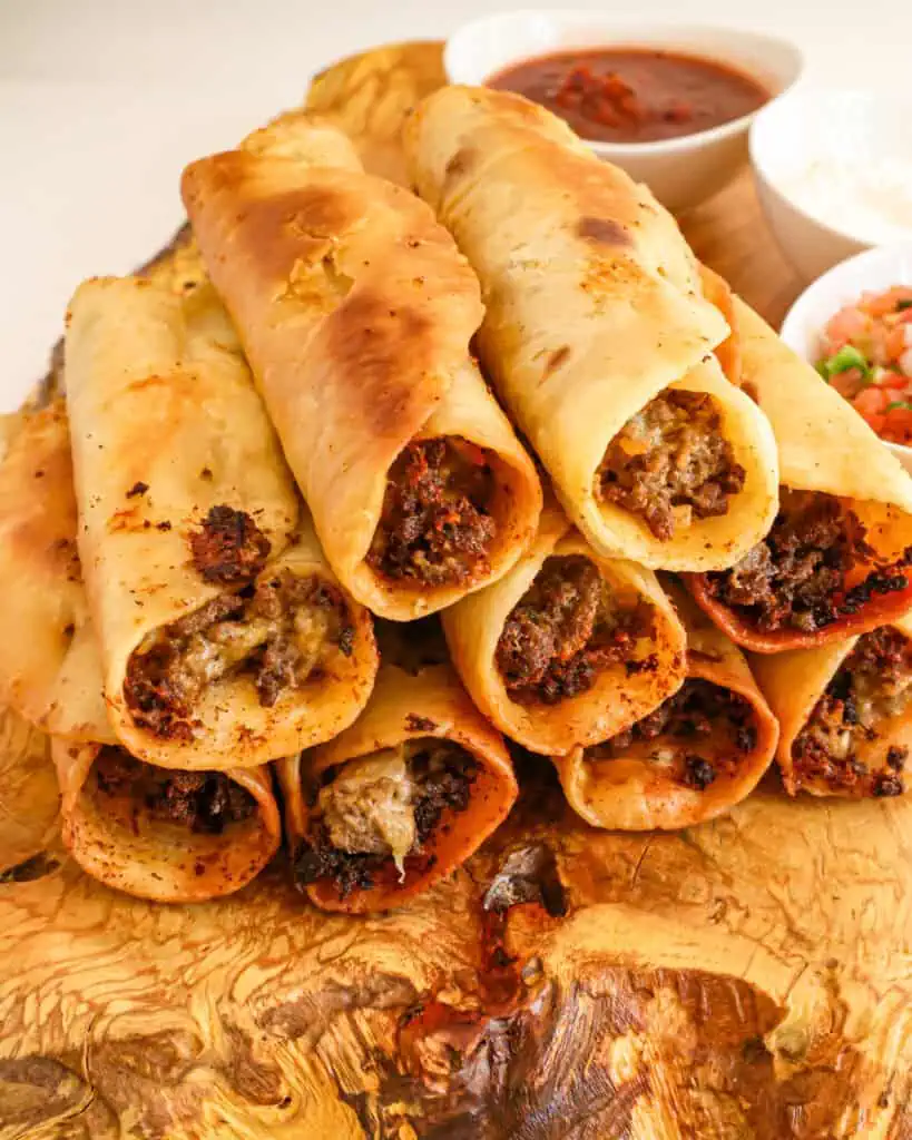 A side view of cheese and beef taquitos stacked on a wooden serving board with small bowl os Pico de Gallo, sour cream, and salsa in the background. 