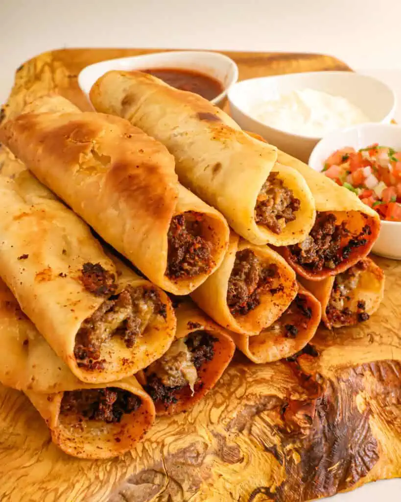 Beef taquitos stacked on a wooden serving board with small white bowls of sour cream, Pico de Gallo, and salsa. 