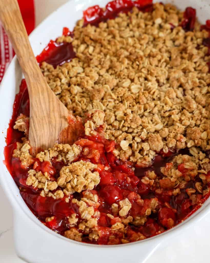 A white oval casserole dish and a wooden spoon full of cherry crumble. 