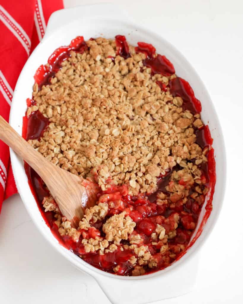 An overhead view of baking dish and a wooden spoon full of cherry crumble. 