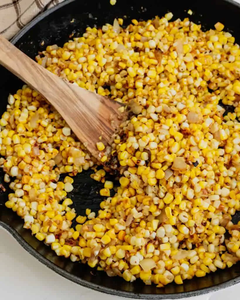 A cast irons skillet and a wooden spatula full of fried corn. 