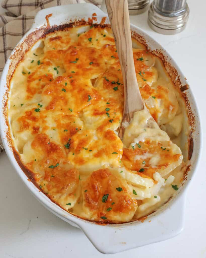 An oval casserole dish full of au gratin potatoes with a wooden spoon in them. 