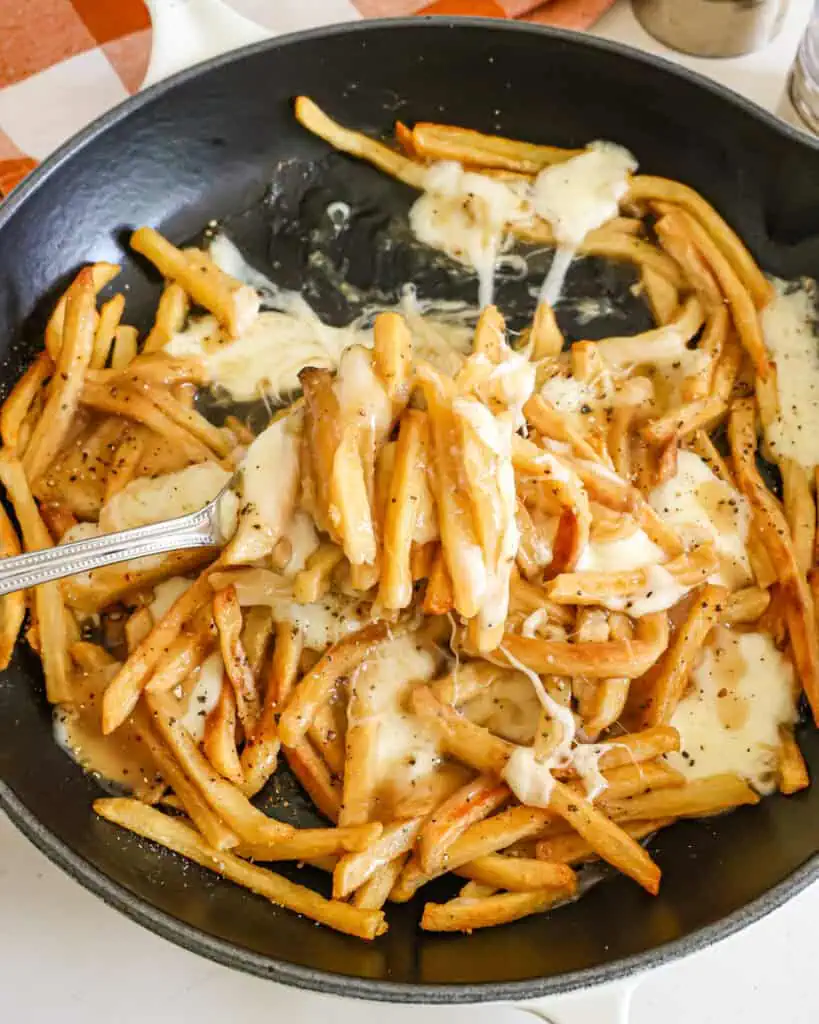 A fork full of french fries with gravy and cheese curbs over a skillet full of them.