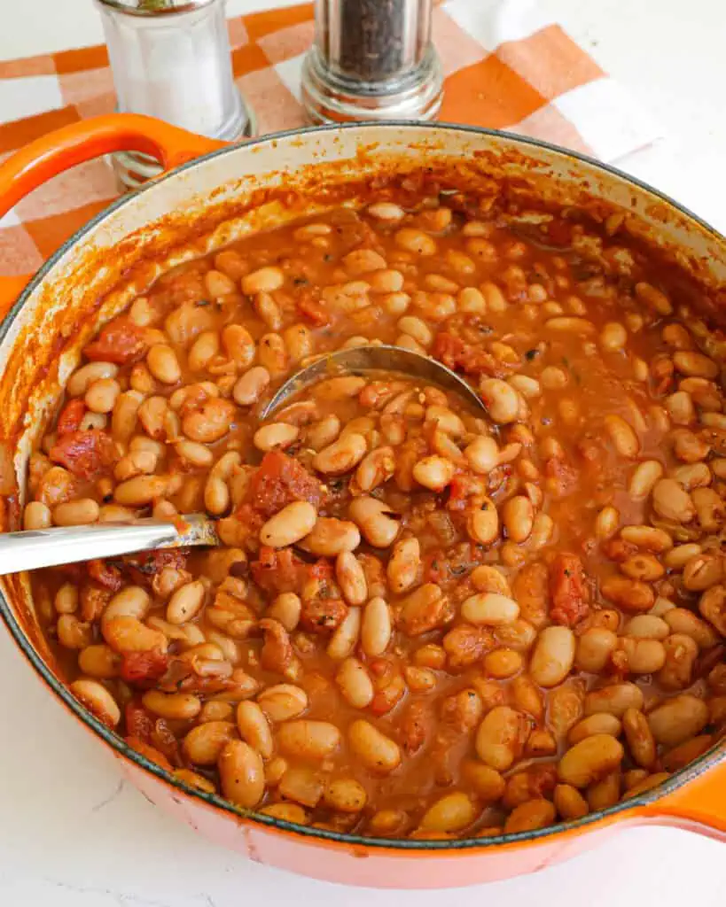 A Dutch oven full of ranch style beans with a ladle in it. There is a salt shaker and pepper mill in the bacckground. 