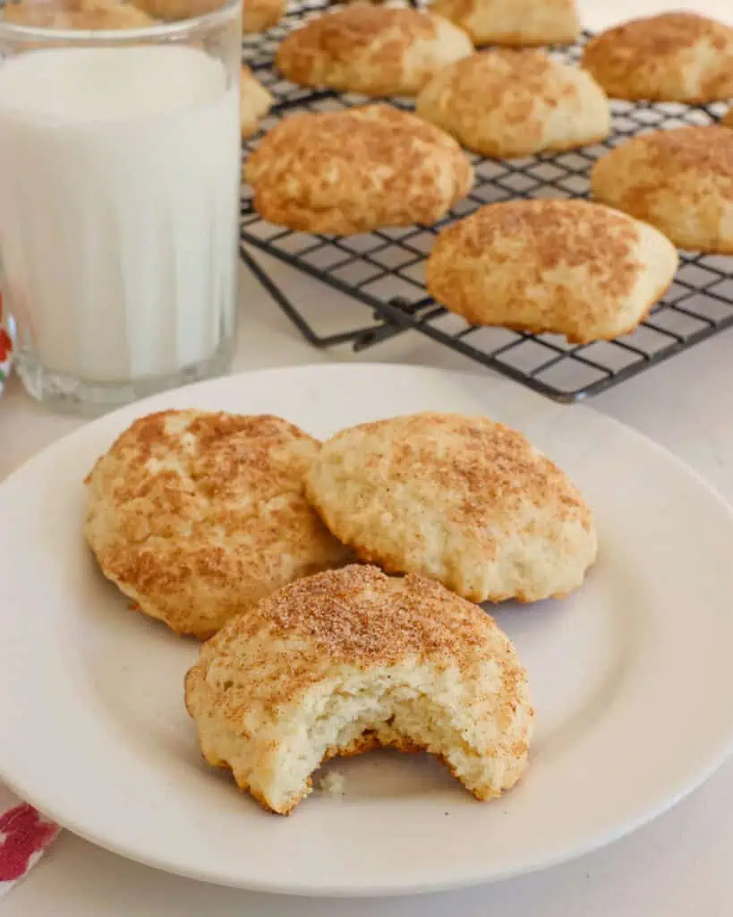Three sour cream cookies on a single serving plate with a glass of milk and a cooling rack full of sour cream cookies. 