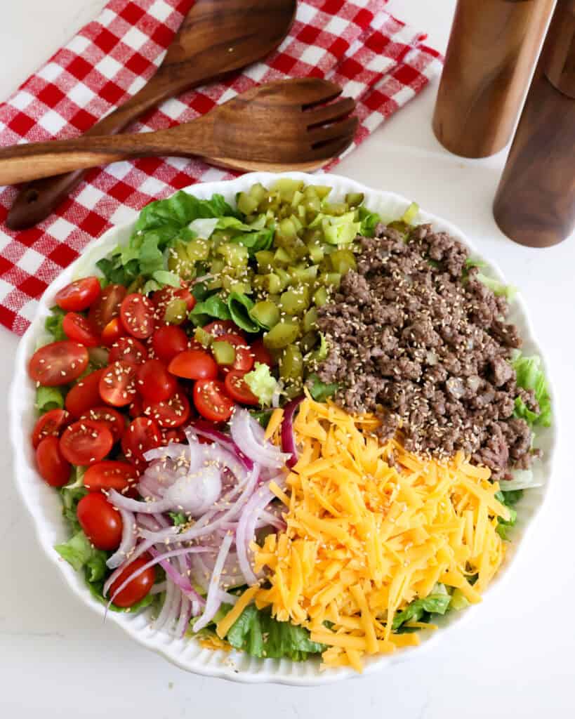 Add the lettuce, ground beef mixture, tomatoes, red onion, cheddar cheese, and dill pickles to a large bowl. Sprinkle with a couple of teaspoons of toasted sesame seeds. 