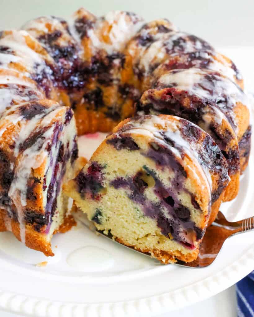 A slice of blueberry cake on a cake server in front of the rest of the cake.