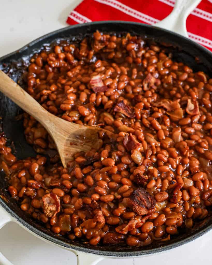 A large cast iron skillet and a large wooden spoon full of baked beans with onions and bacon. 