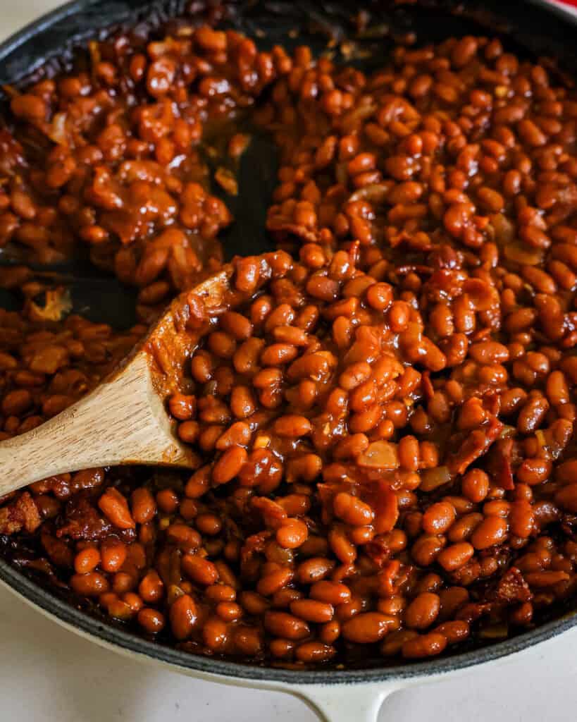 A close up view of baked beans on a wooden spoon in a cast iron skillet full of baked beans. 