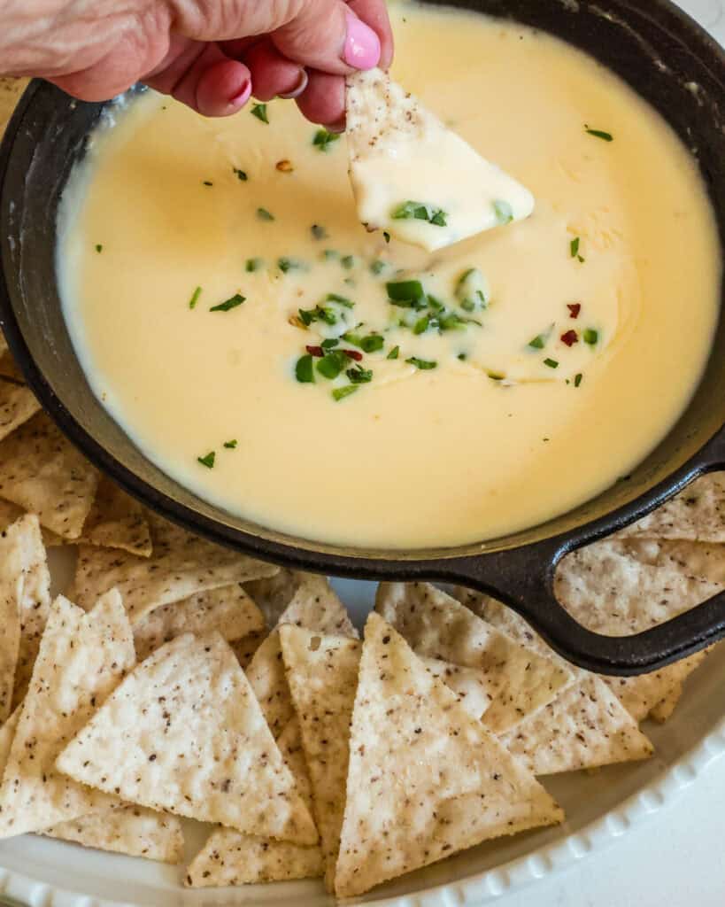 A tortilla chips dipped in cheese dip above a small cast iron skillet full of cheese dip. 