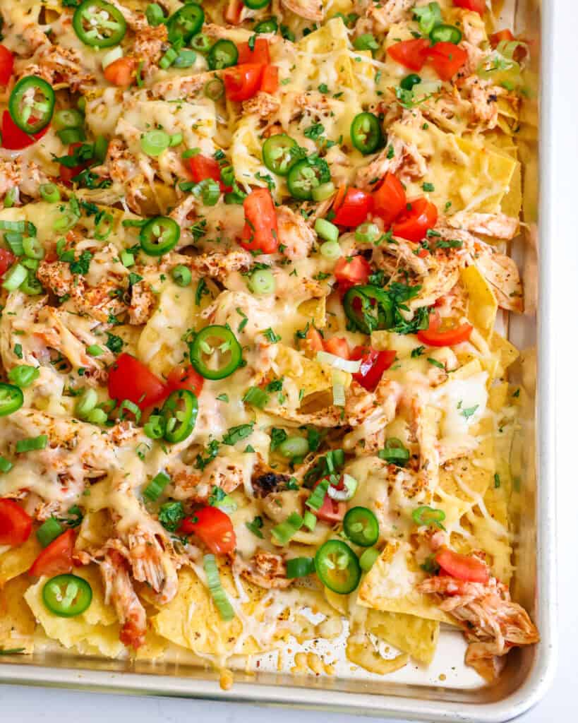 A parchment paper-lined baking sheet full of chicken nachos topped with chopped tomatoes, green onions, jalapeno, and cilantro. 