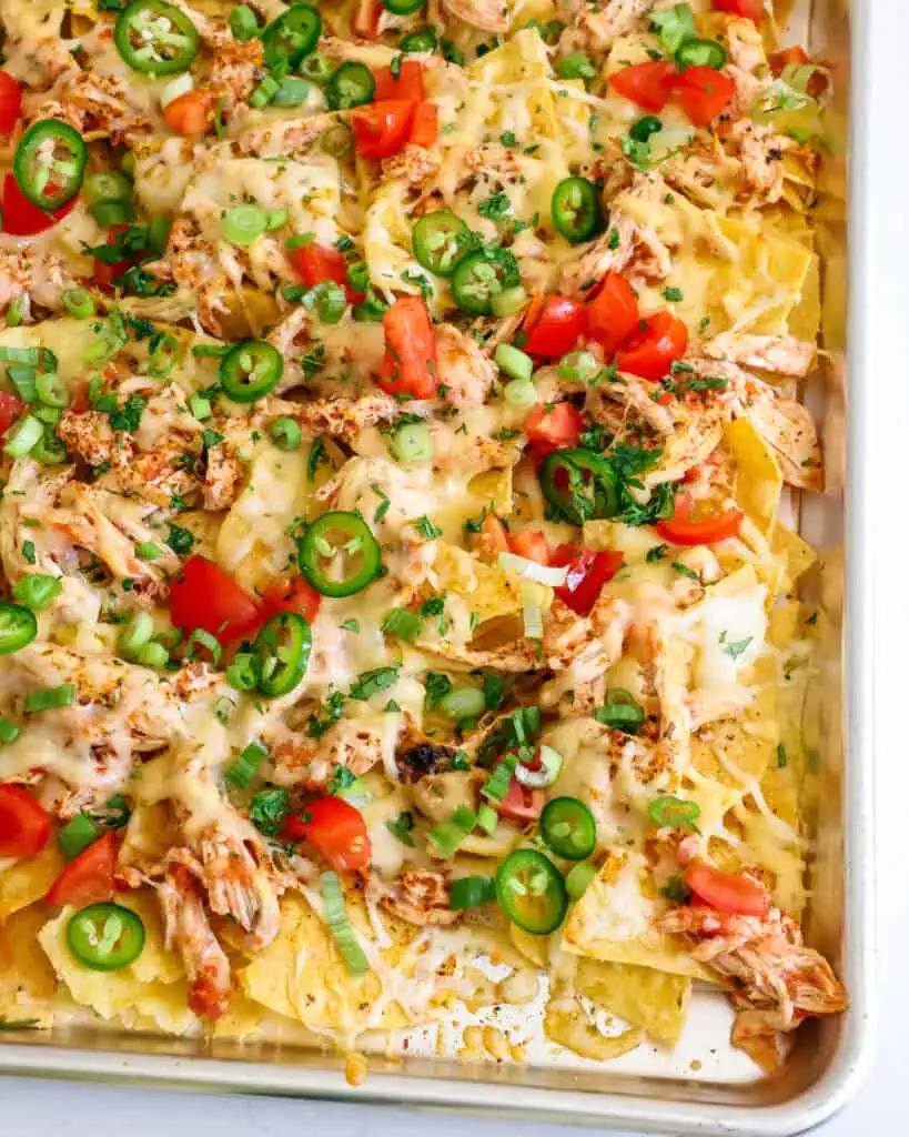 A parchment paper-lined baking sheet full of chicken nachos topped with chopped tomatoes, green onions, jalapeno, and cilantro. 