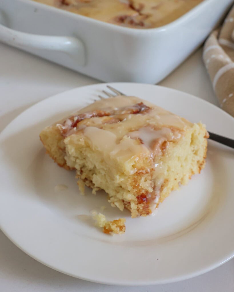 A piece of cinnamon roll cake on a plate with a fork in front of the baking dish with the cake in it. 