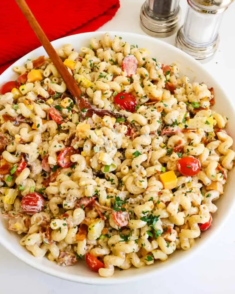 A serving bowl and a wooden spoon full of cowboy pasta salad with a salt and pepper shaker in the background. 