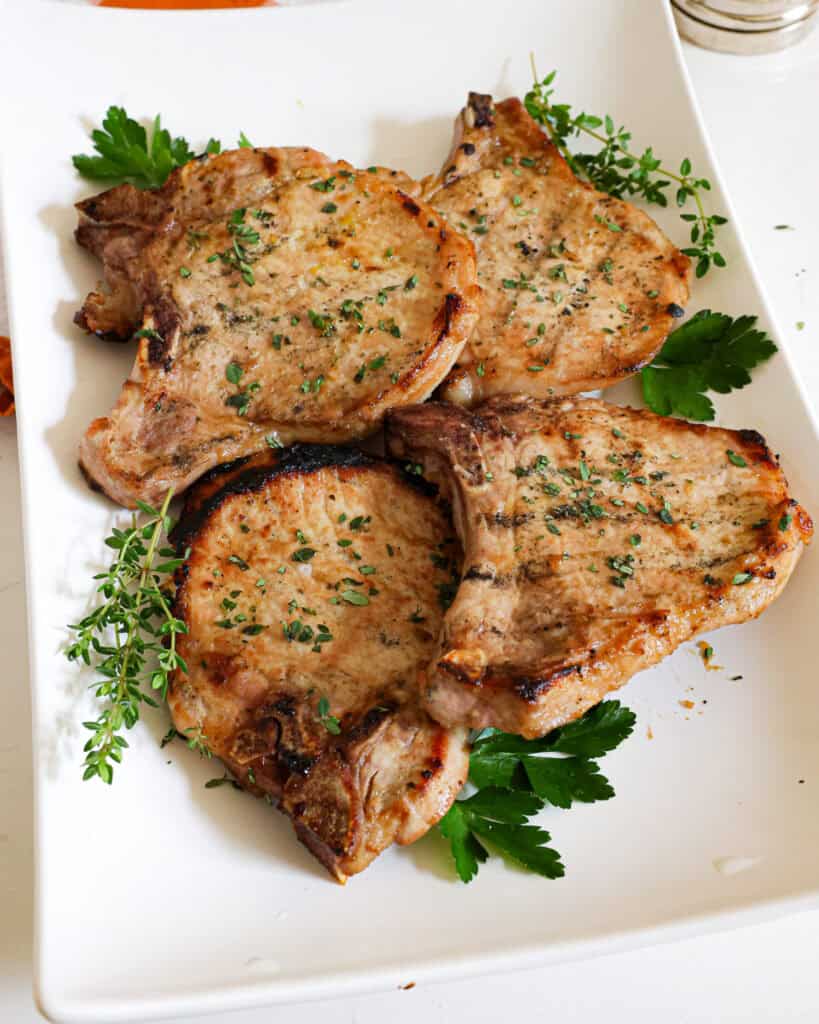Four grilled pork chops garnished with chopped thyme and parsley on a white serving platter.