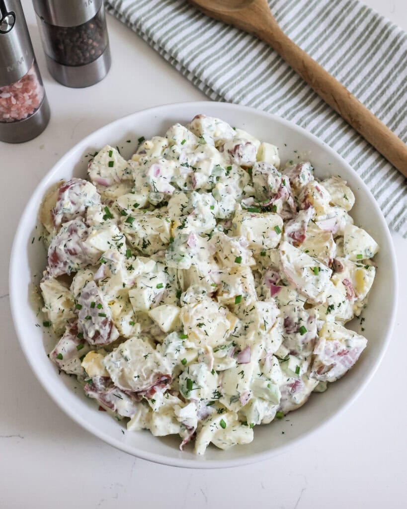An overhead view of red potato salad in a white serving bowl with a wooden spoon and salt and pepper shaker in the background. 
