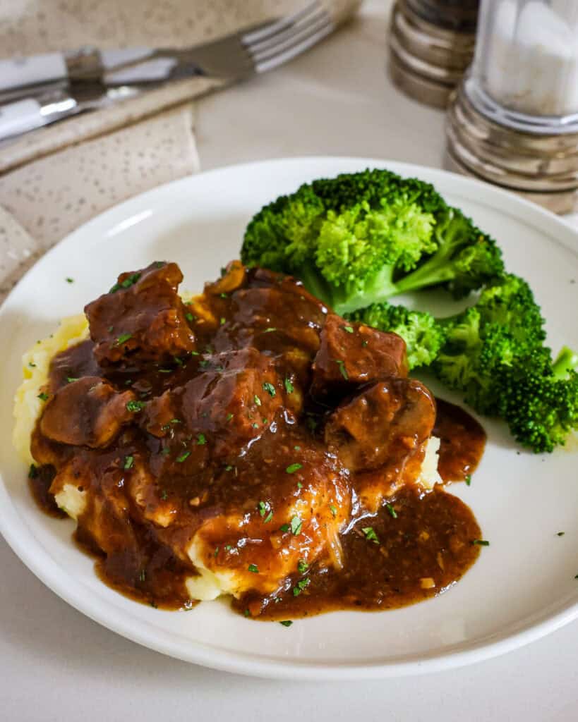 Beef tips and gravy over mashed potatoes with a side of steamed broccoli on a single serving white plate. 