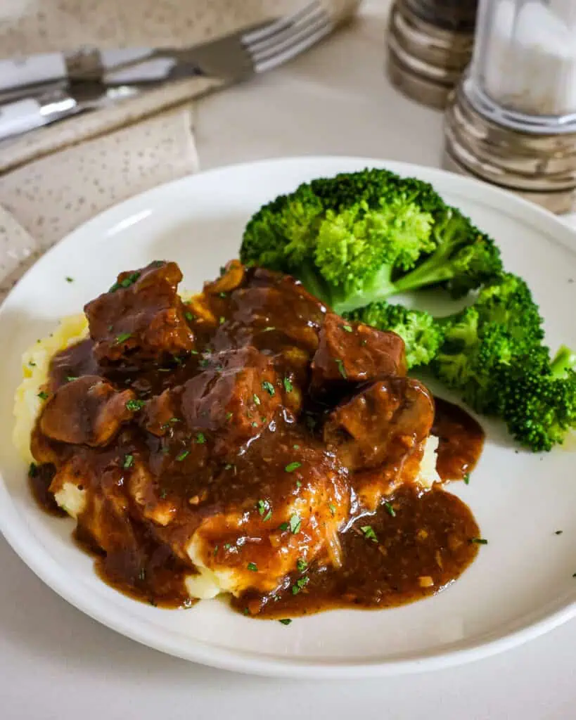 Beef tips and gravy over mashed potatoes with a side of steamed broccoli on a single serving white plate. 