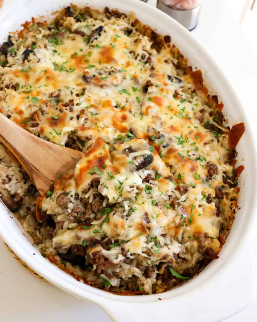 A white oblong casserole full of ground beef and cheese casserole with onions and mushrooms. 