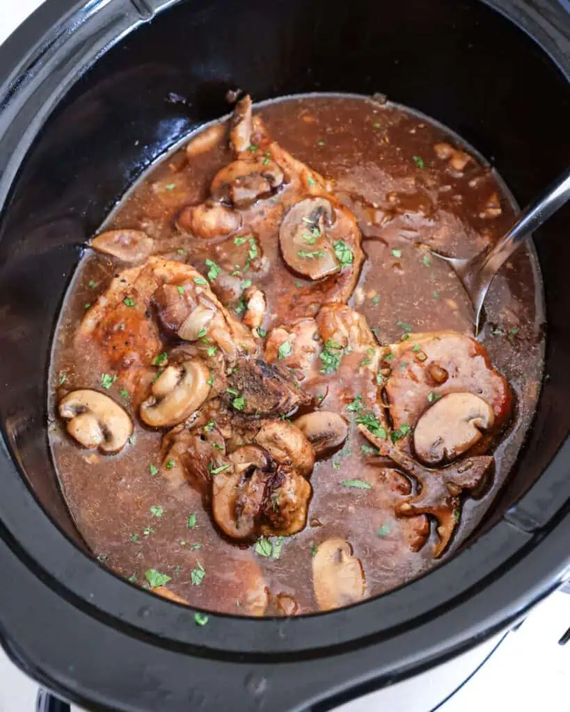 A large black crock pot full pork chops, onions, mushrooms, and gravy.