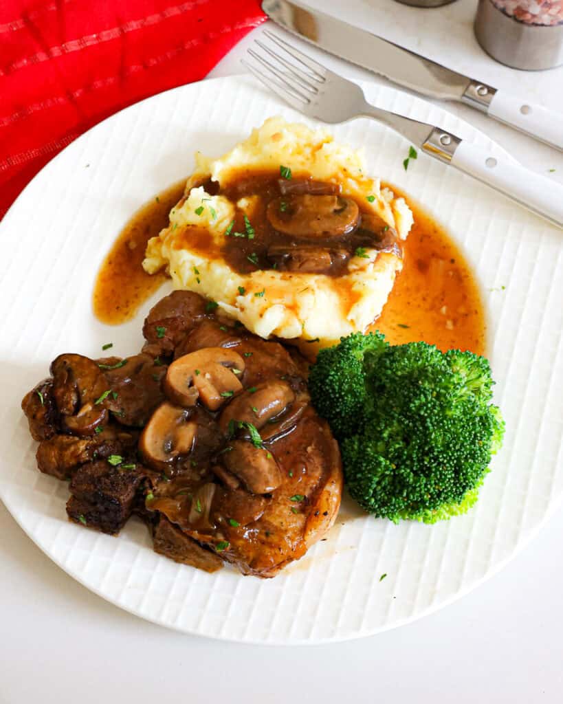 An overhead view of a white serving plate with a crock pot pork chop, mashed potatoes and gravy, and steamed broccoli. 