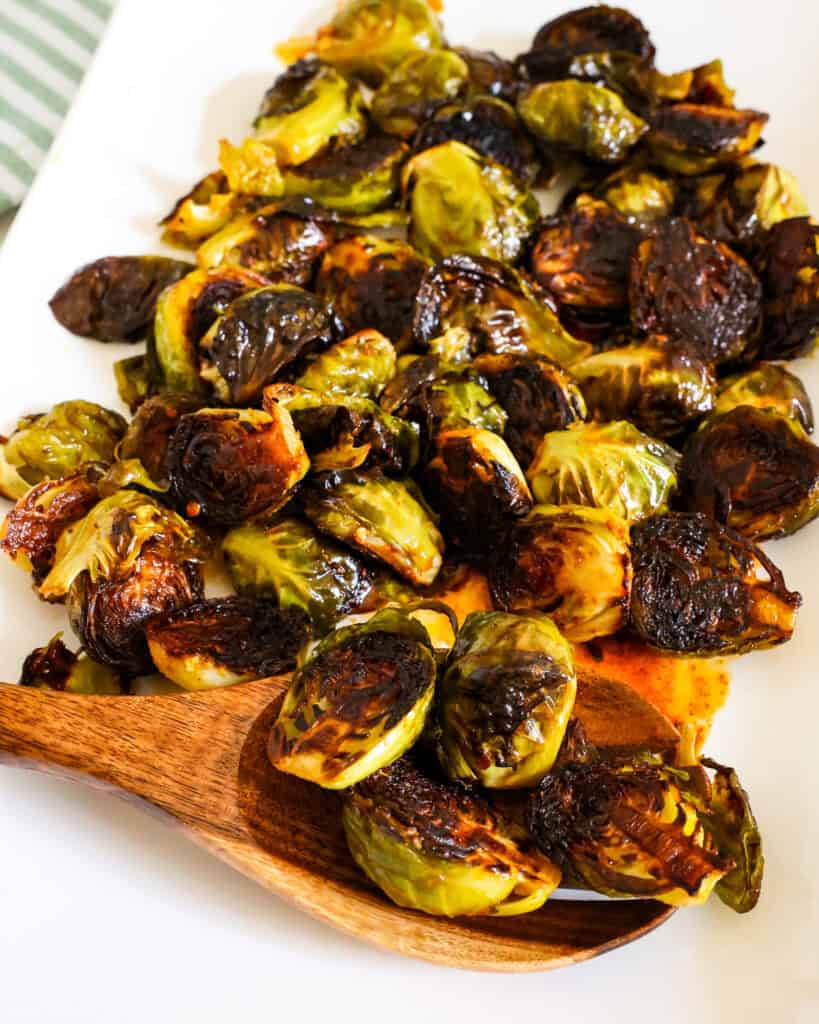A white serving platter and a wooden spoon full of crispy Brussels sprouts.