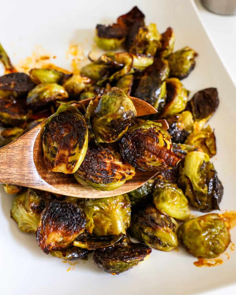 A close up view of crispy Brussels sprouts in a wooden spoon over a platter full of them.