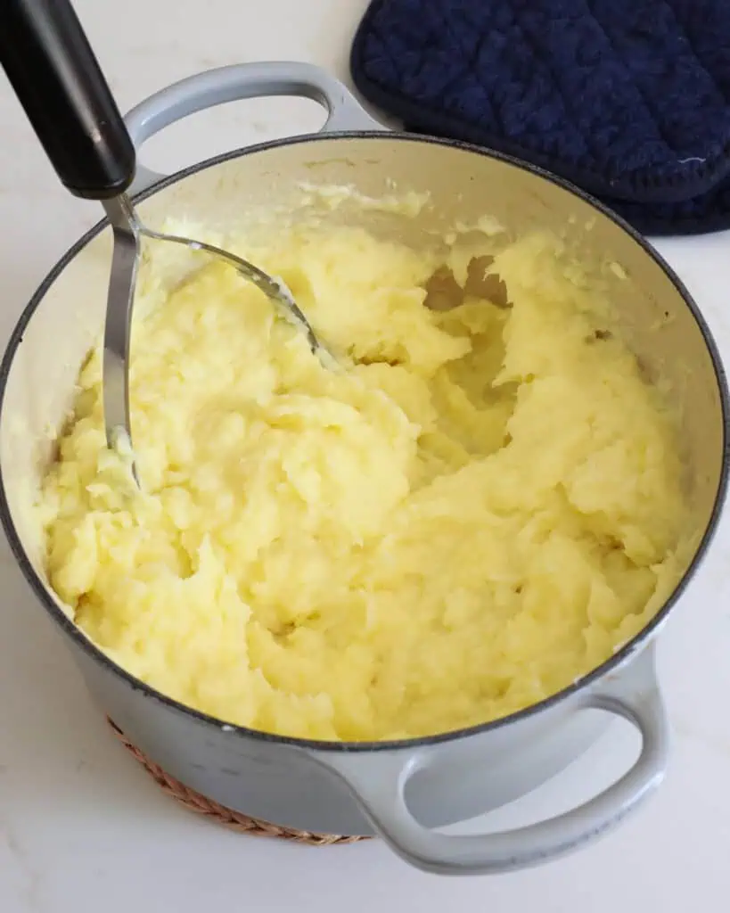 A Dutch oven full of mashed potatoes with a potato masher in the pot.