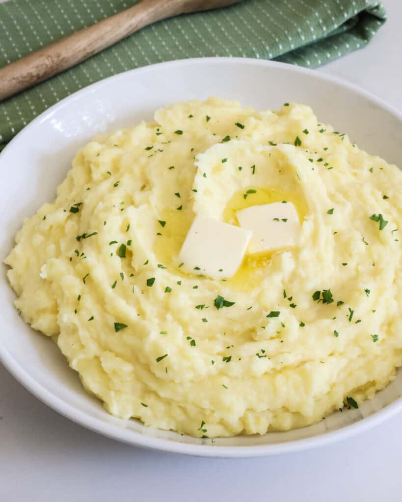 A white serving bowl full of mashed potatoes garnished with chopped parsley. 