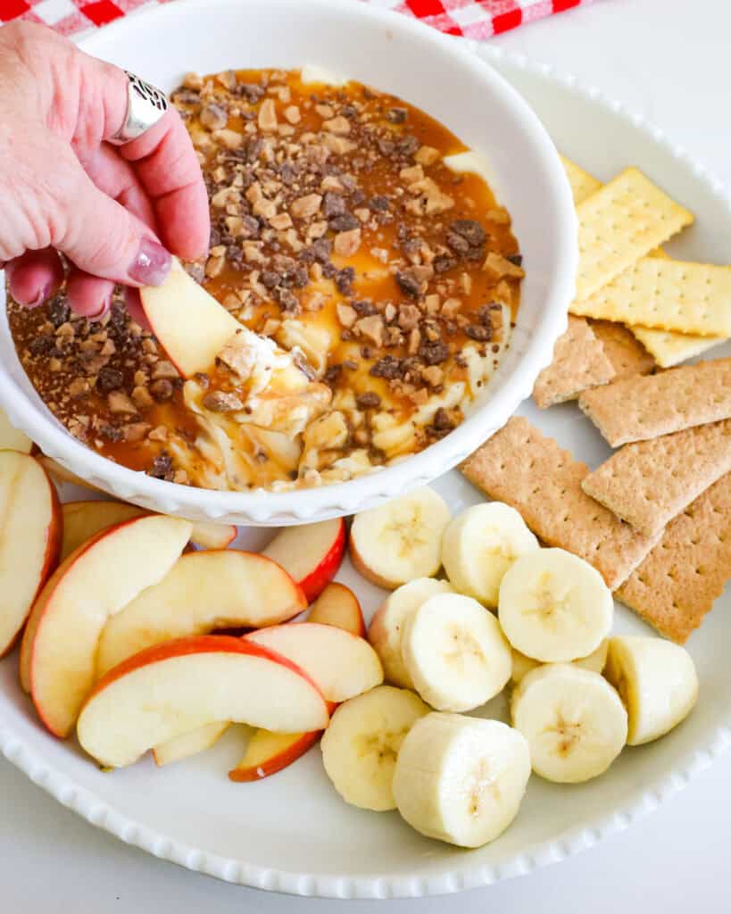 A serving bowl full of caramel apple dip on a platter with apple slices, banana slices, graham crackers. and Club crackers. 