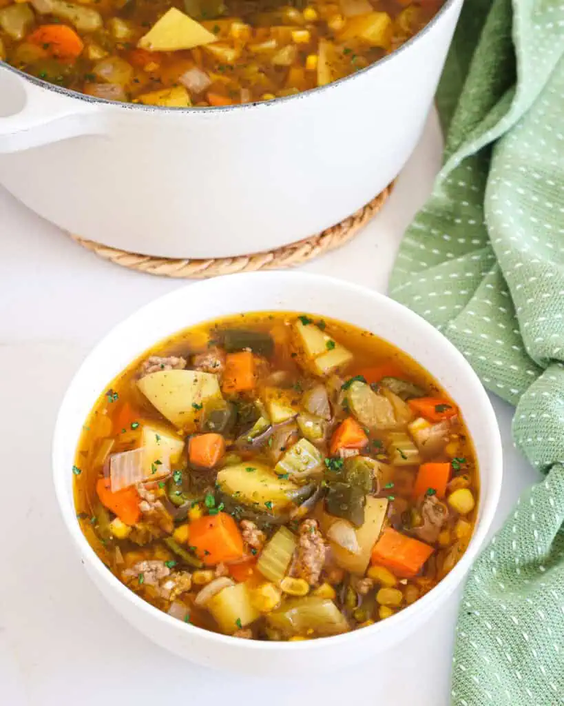 A single serving white bowl full of hamburger potato soup with a Dutch oven full of it in the background. 