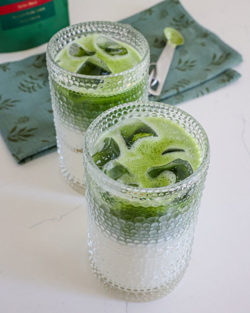 Two glass of milk with matcha mixture added to them. There is a green towel, measuring spoons, and matcha powder in the background. 