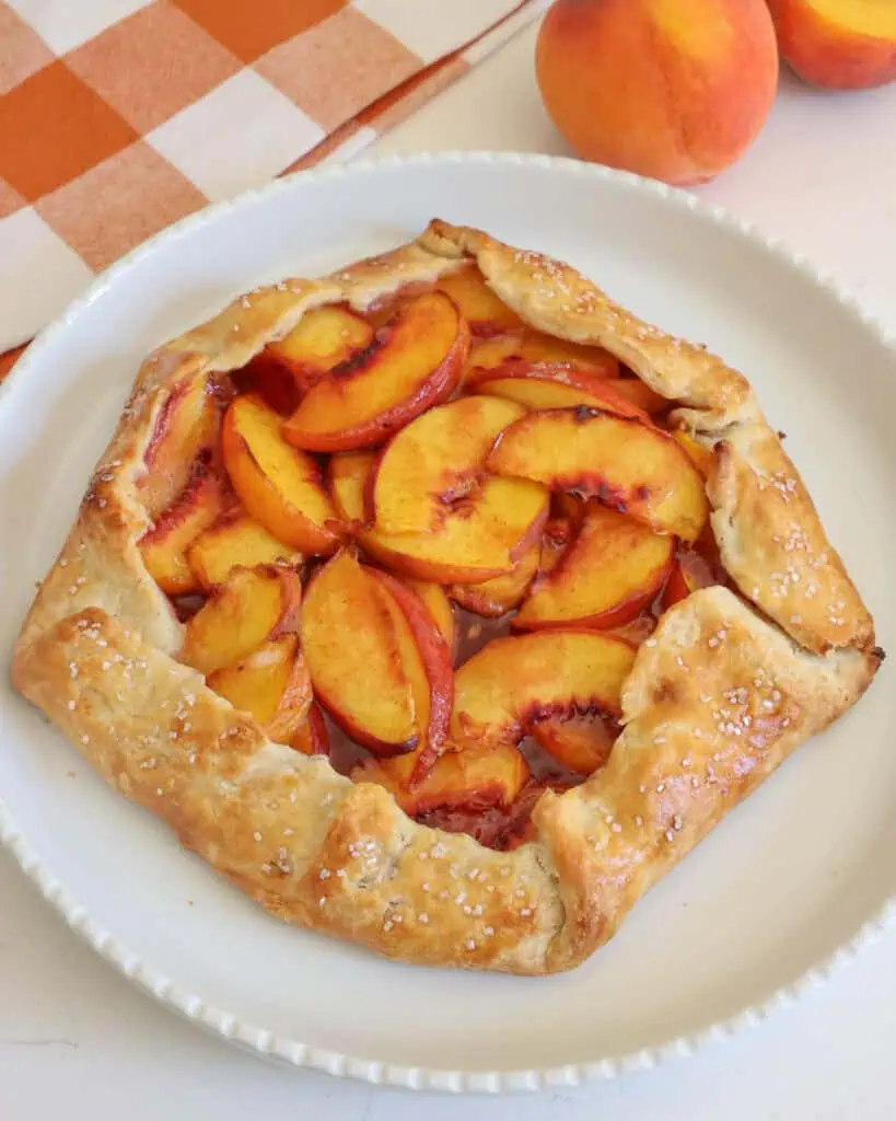 A peach galette on a white serving platter with peaches and a kitchen towel in the background.