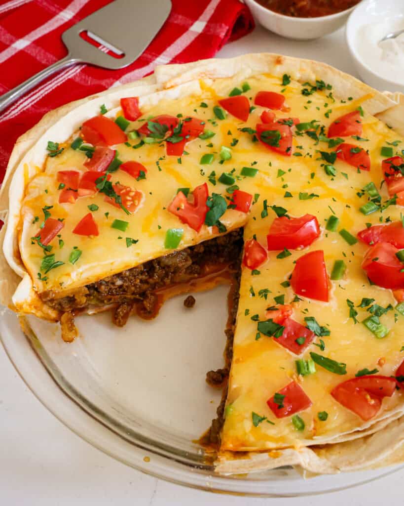 A taco pie with a slice cut out of it. A red towl and pie server are in the background.