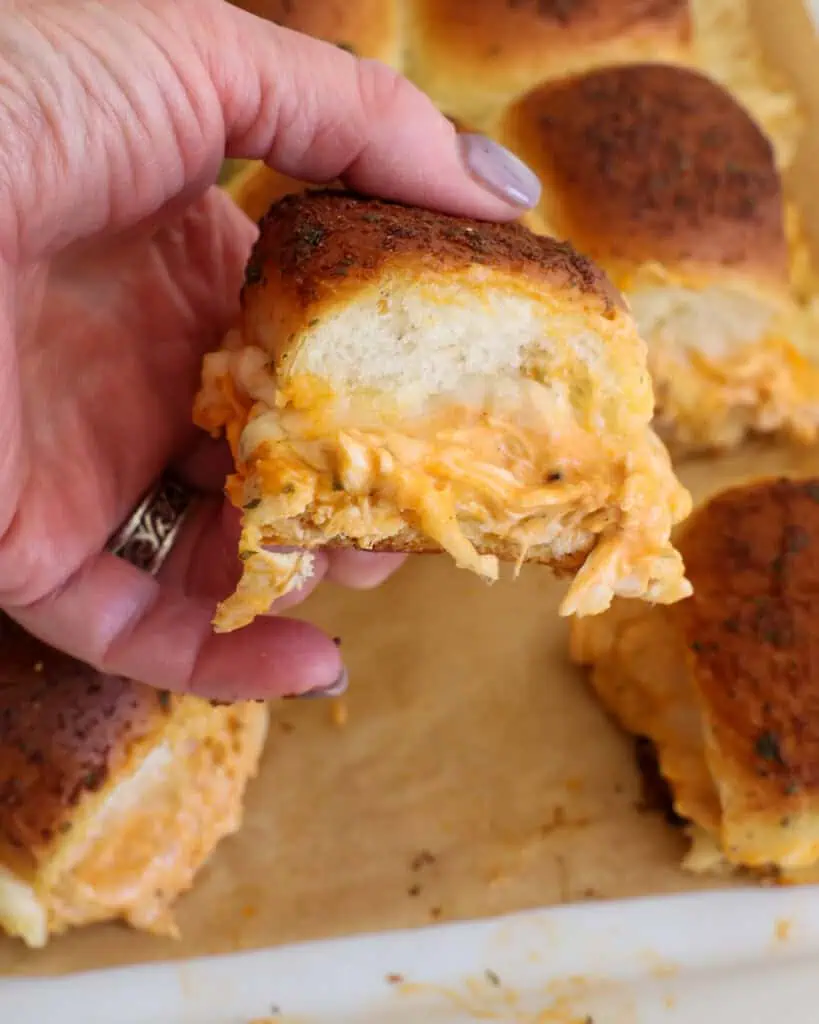 A woman's hand holding a buffalo chicken slider over a baking sheet full of buffalo chicken sliders.