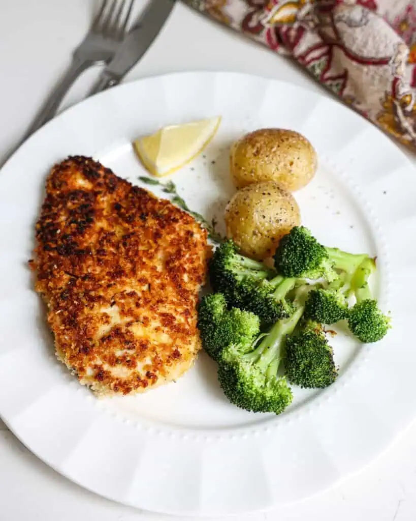 Crispy breaded chicken, broccoli, and baby potatoes on a single serving plate with a napkin and fork and knife in the background.