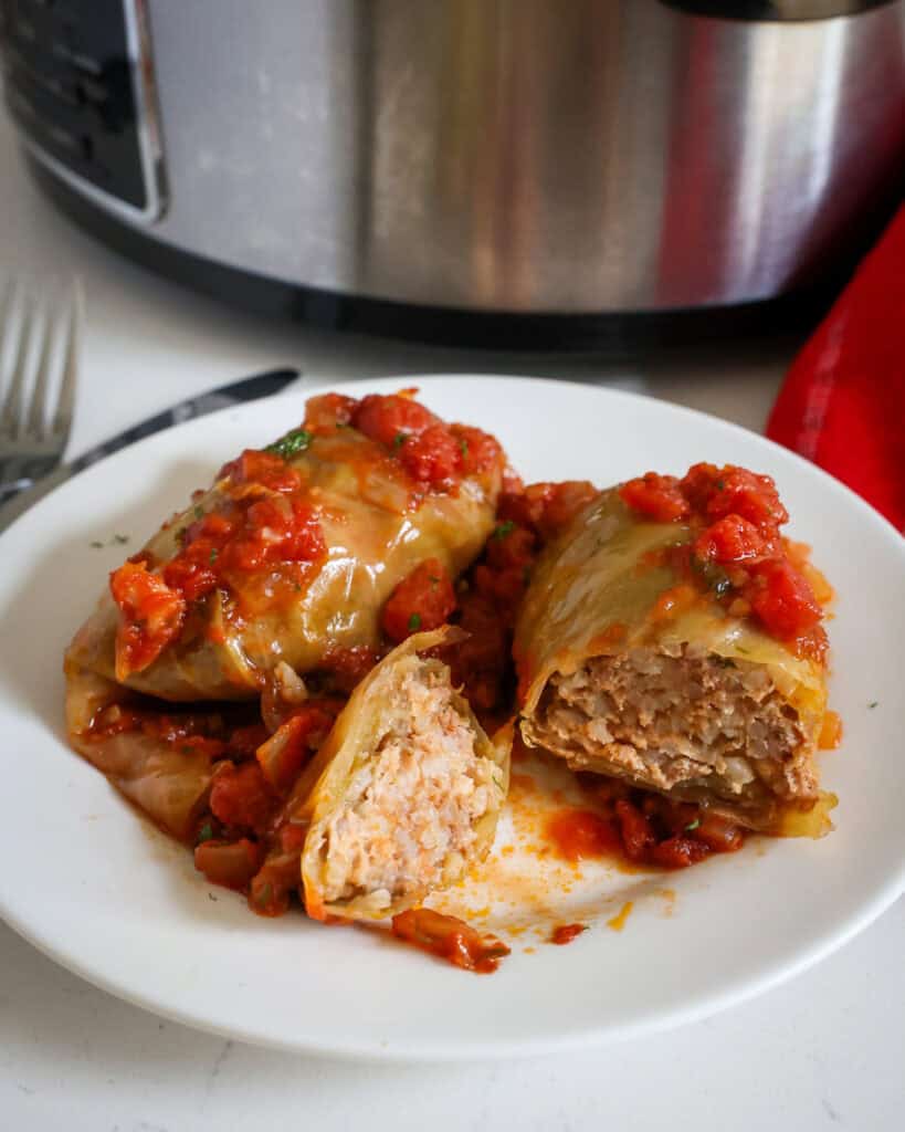 A sliced open cabbage roll on a single serving plate with another cabbage roll. A fork and knife and crockpot are in the background. 