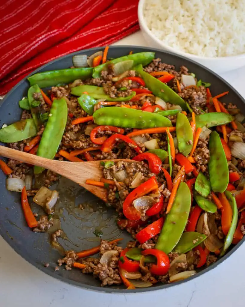 A wooden spoon and a large black skillet full of ground beef stir fry with mixed vegetables.