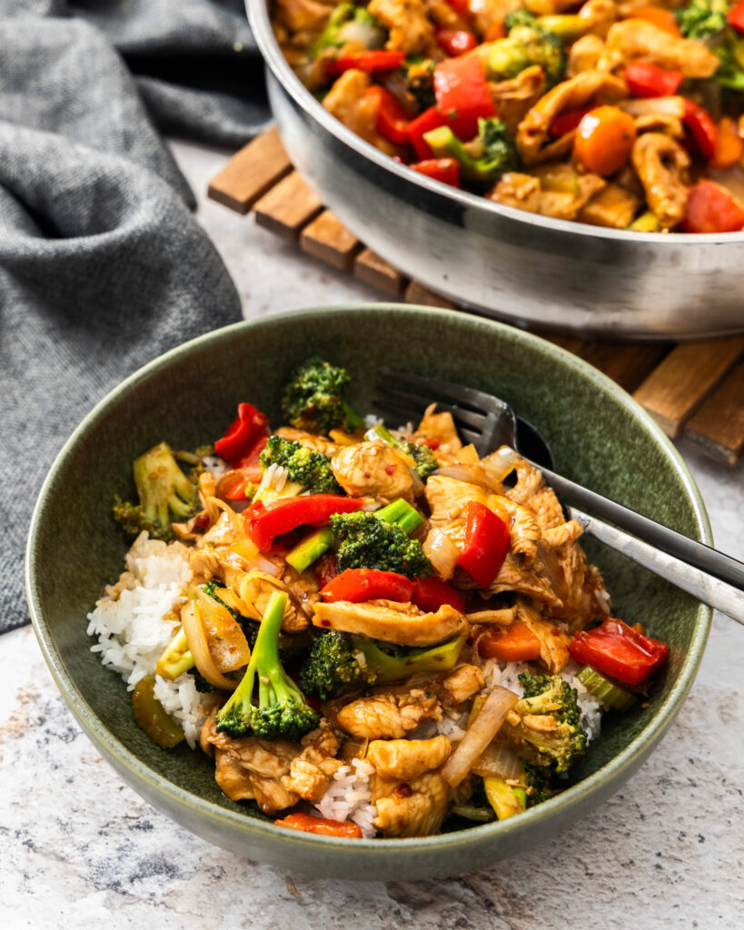 Hunan chicken in a bowl over rice with a skillet full of more hunan chicken in the background.