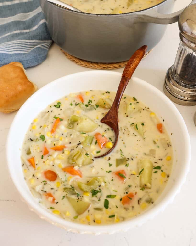 A single serving white bowl full of potato corn chowder. There is a roll and a salt and pepper shaker in the background.