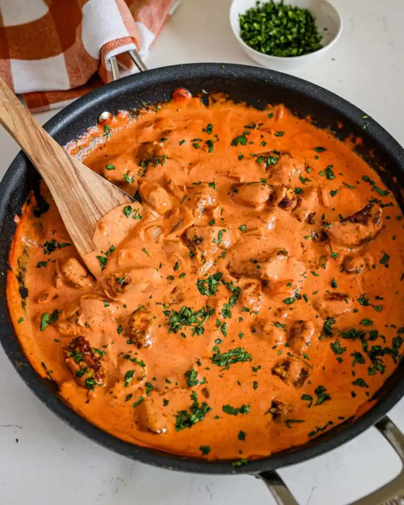 A skillet and wooden spatula full of butter chicken garnished with chopped cilantro.